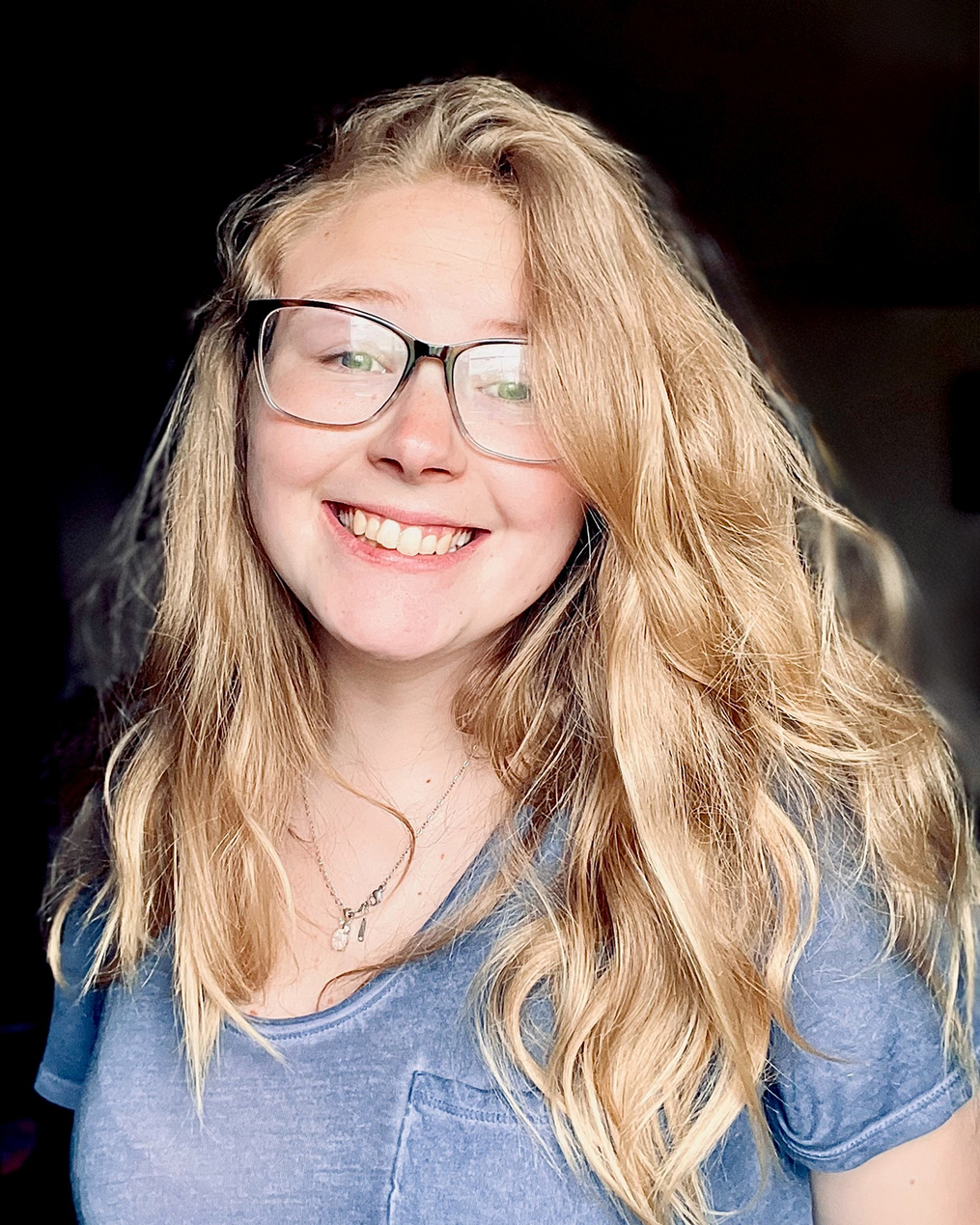 Smiling young woman with long blonde hair and glasses, wearing a blue T-shirt, photographed indoors against a dark background.